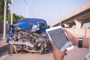 Man using tablet to file insurance claim after severe car accident, showing front-end vehicle damage and roadside scene.
